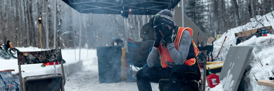 Person wearing mittens and winter gear cover their face while sitting at a chair at a snowy site in the woods