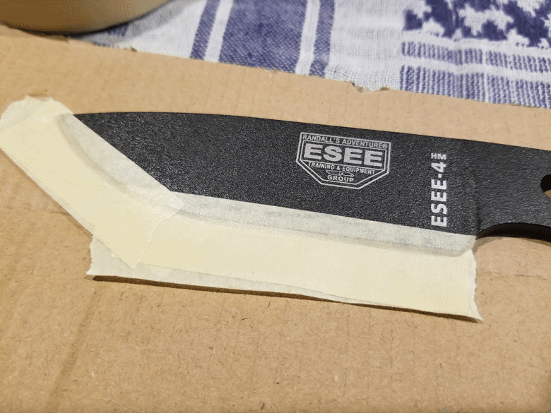 Black powder coated knife blade taped to a piece of cardboard with masking tape