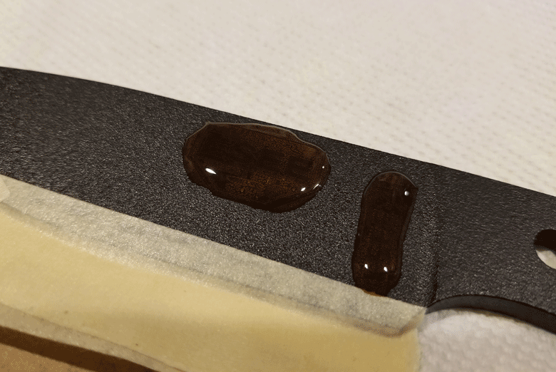 Portion of black powder coated knife blade with drops of acid on it and  taped to a white mat working surface